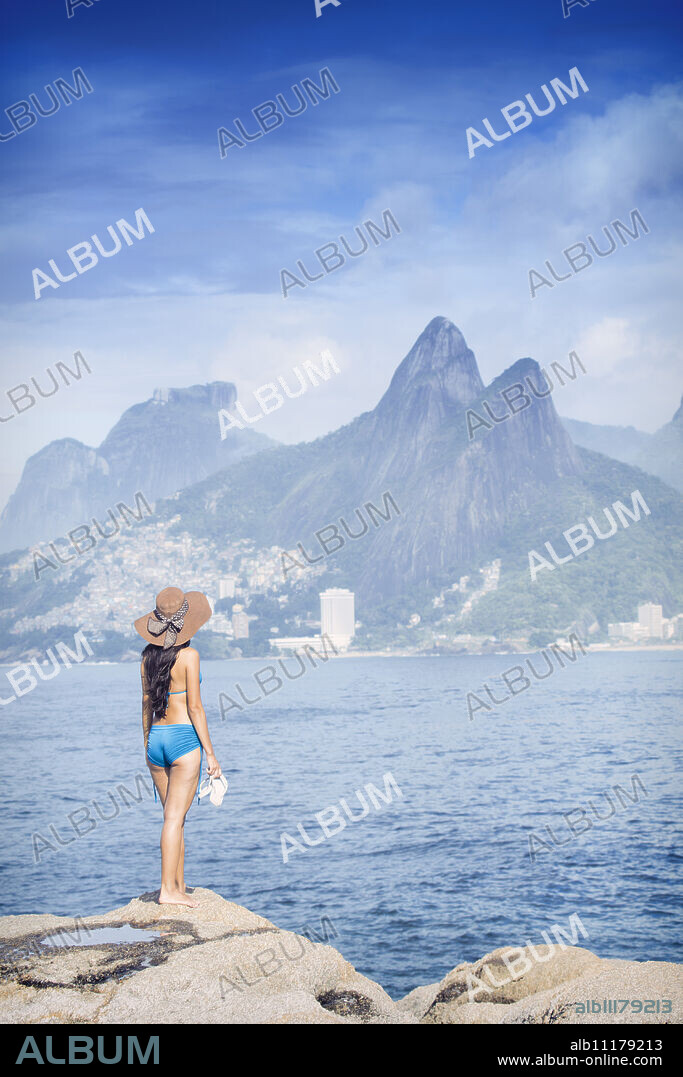 A 20-25 year old young Brazilian woman standing on the Arpoador rocks with Ipanema and the Morro Dois Irmaos hills in the distance, Rio de Janeiro, Brazil, South America.