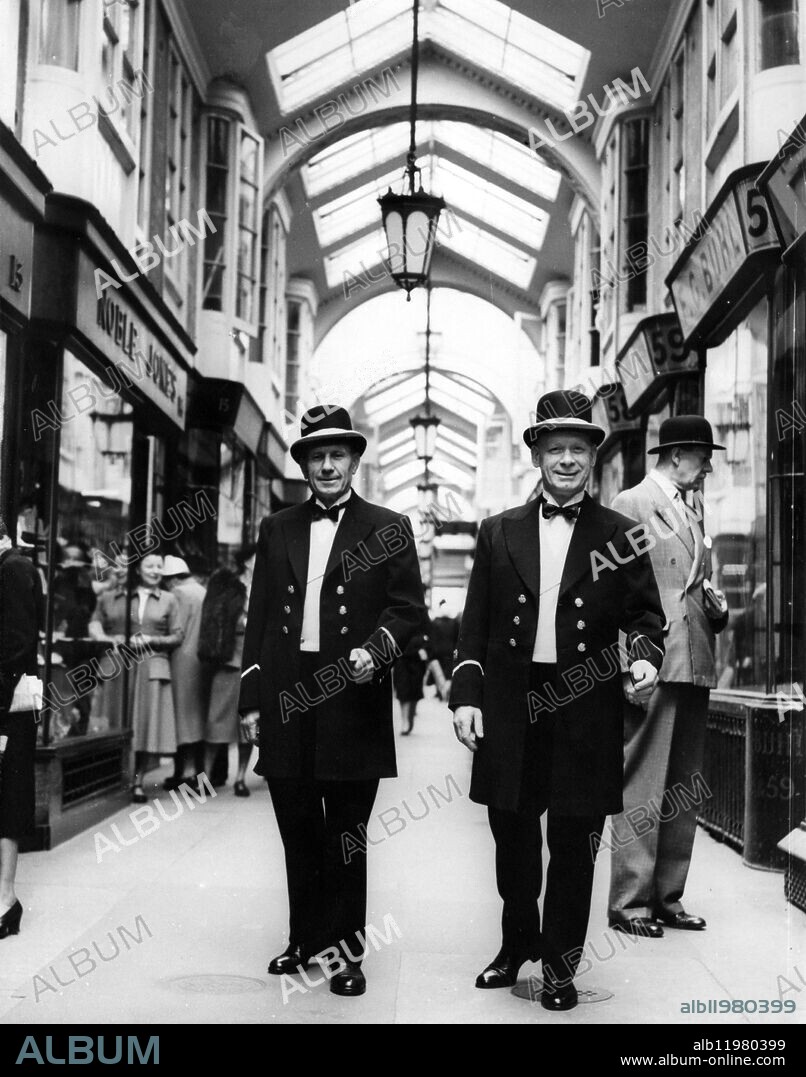 Uniforms for Burlington Arcade Beadles . The two Beadles who , each day at 5.30 pm , ring the 100 year old bell for closing time in the famous Burlington Arcade , have just received new uniforms which they will wear during the summer . Made by Sam Fisher , a tailor in the Arcade , using old prints for guidance , they consist of black doeskin frock coat , yellow , double - breasted waistcoat and pure silk top hat . Photo shows ; The two Beadles wearing their new uniforms for the first time walking in the Burlington Arcade . They are , ( left ) Mr Frank Clark of Fulham and Chief Beadle , Robert Baker of Stockwell . 29 May 1952.