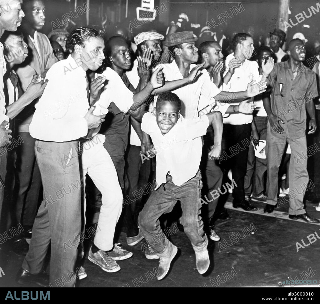 Entitled: "Part of crowd in Harlem chants and taunts police on Lenox Avenue last night." On Thursday, July 16, 1964, James Powell was shot and killed by police Lieutenant Thomas Gilligan. The second bullet of three fired by Lieutenant Gilligan killed the 15-year-old African American in front of his friends and about a dozen other witnesses. The incident immediately rallied about 300 students from a nearby school who were informed by the principal. This incident set off six consecutive nights of rioting that affected the New York City neighborhoods of Harlem and Bedford-Stuyvesant. In total, 4,000 New Yorkers participated in the riots which led to attacks on the New York City Police Department, vandalism, and looting in stores. At the end of the conflict, reports counted one dead rioter, 118 injured, and 465 arrested. Photographed for the New York World-Telegram & Sun by Stanley Wolfson, 1964.