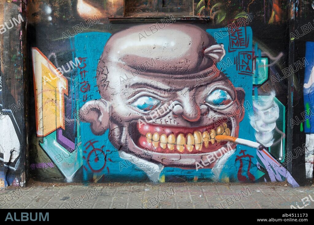 Caricatura de cabeza calva con ojos saltones mostrando la dentadura que sostiene un cigarrillo, graffiti mural, calle Selva de Mar, barrio Poblenou, Barcelona en 2014.