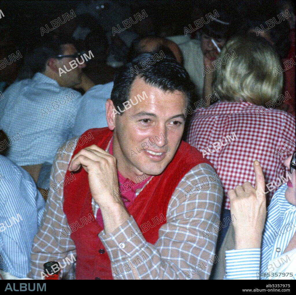 ROCK HUDSON. Circa 1957, Los Angeles, California, USA: American actor ROCK HUDSON in red waistcoat enjoying dinner with friends at a restaurant in Hollywood.