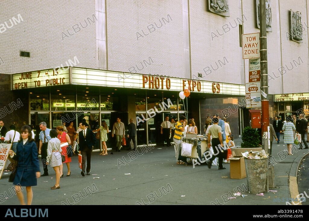 View of people walking outside of the New York Coliseum convention center, at Columbus Circle and 58th Street, on the west side of Manhattan, New York City, June, 1969. Marquee signs at the entrance advertise the 1969 Photo Expo taking place from June 7 to 15.
