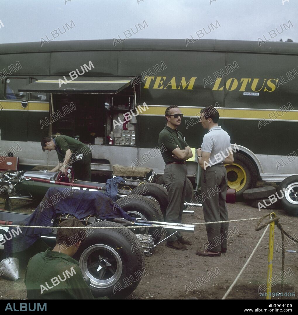Chapman and Clark outside the Lotus team bus, French Grand Prix, Clermont-Ferrand, France, 1965. Colin Chapman is wearing sunglasses and listening to Jim Clark speak. To the left of them two mechanics work on a car. Chapman was the founder of Lotus cars and Clark was one of his best drivers. Clark won the French Grand Prix for Lotus in a Lotus Climax.