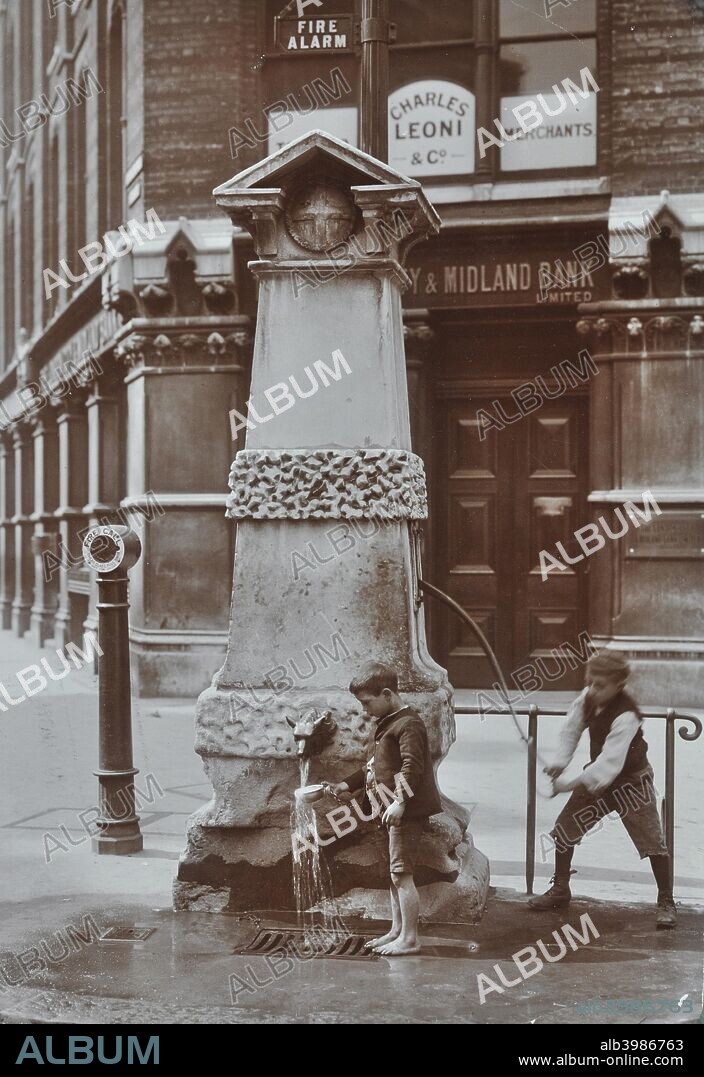 Children drawing water from the Aldgate pump, London, August 1908. One boy pumps while the other holds a cup under the water outlet. To the left is a street fire alarm, with a bank behind.