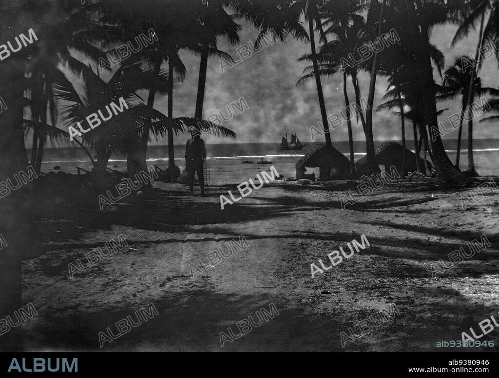 Atiu, Mauke or Aitutaki, William Saunderson Cooper, 1910s, Southern Cook Islands, View of the outside of a number of huts/houses, set among palm trees. The lagoon is visible in the background, and a small ship is beyond the reef.