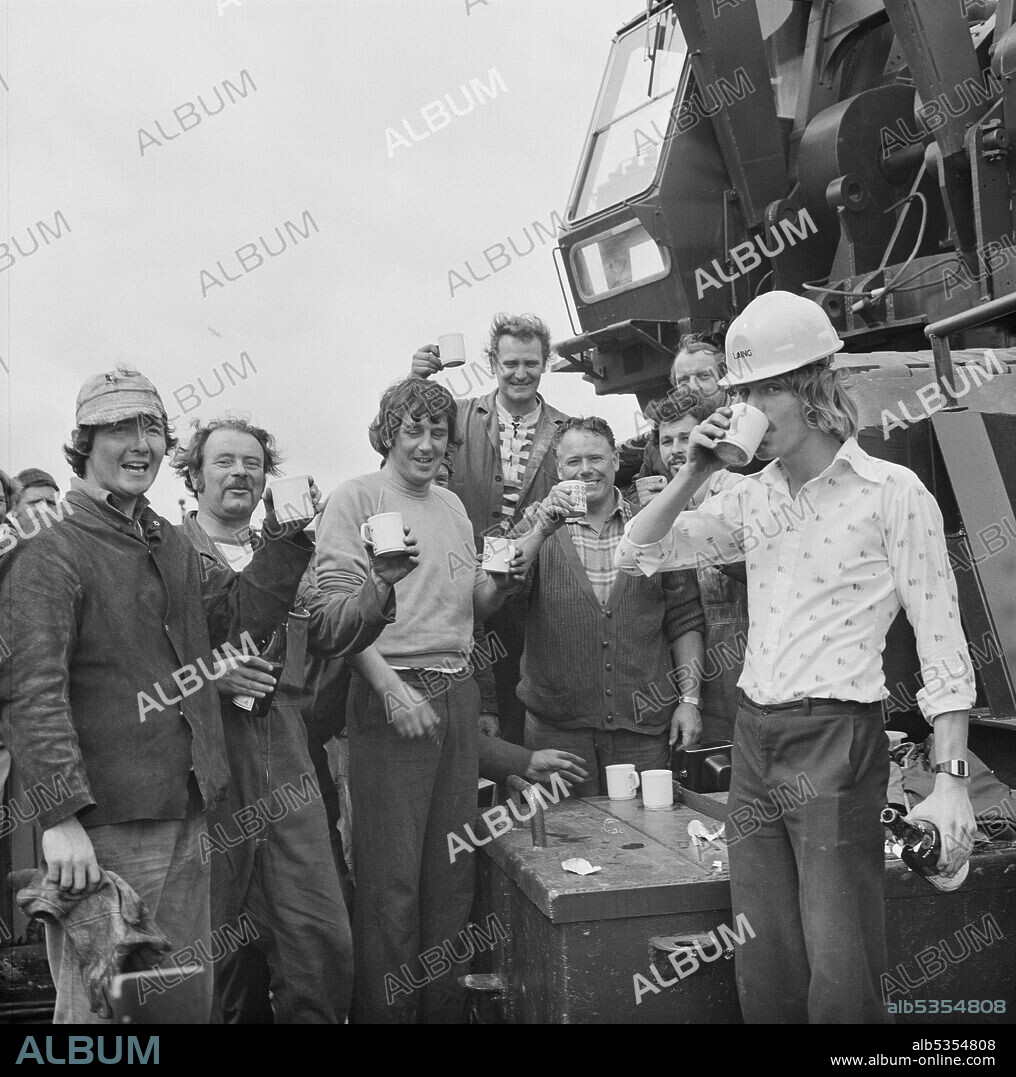 JOHN LAING PLC. A group of workers toasting the launch of a twin barge from Woolston, Hampshire, for use in the construction of the Peel Common Sewage Works outfall at Browndown. Laing Civil Engineering were contracted to construct the Browndown Outfall for the Southern Water Authority, as part of the South Hampshire Main Drainage Scheme for the treatment and disposal of sewage. Browndown Outfall is a 1km long underwater culvert which transports treated sewage from Peel Common Sewage Works, another contract carried out by Laing. The outfall was built in sections which were linked together in a chain on the seabed. The landward section of the outfall was cast in-situ in a temporary cofferdam.