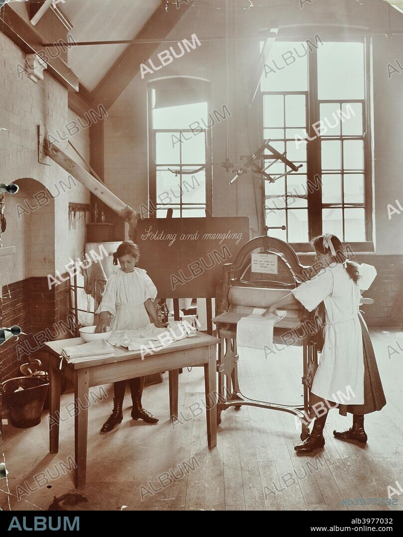 Laundry work, Tennyson Street School, Battersea, London, 1907. One girl folds clothes on a table while another puts them through a mangle. Behind them a blackboard reads; 'Folding and mangling'.