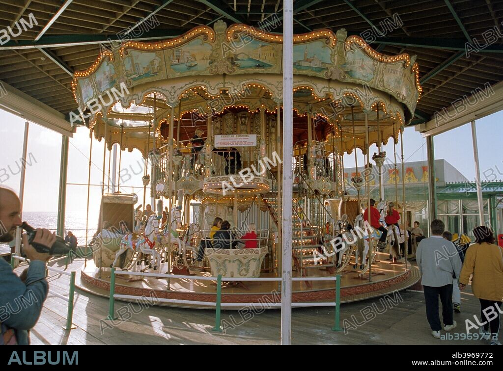 Venetian Carousel, North Pier, Blackpool, Lancashire, 1999. Children ride around on this unusual two storey carousel. In the background is the sea while on the left is a parent or tourist filming from a hand held camera.