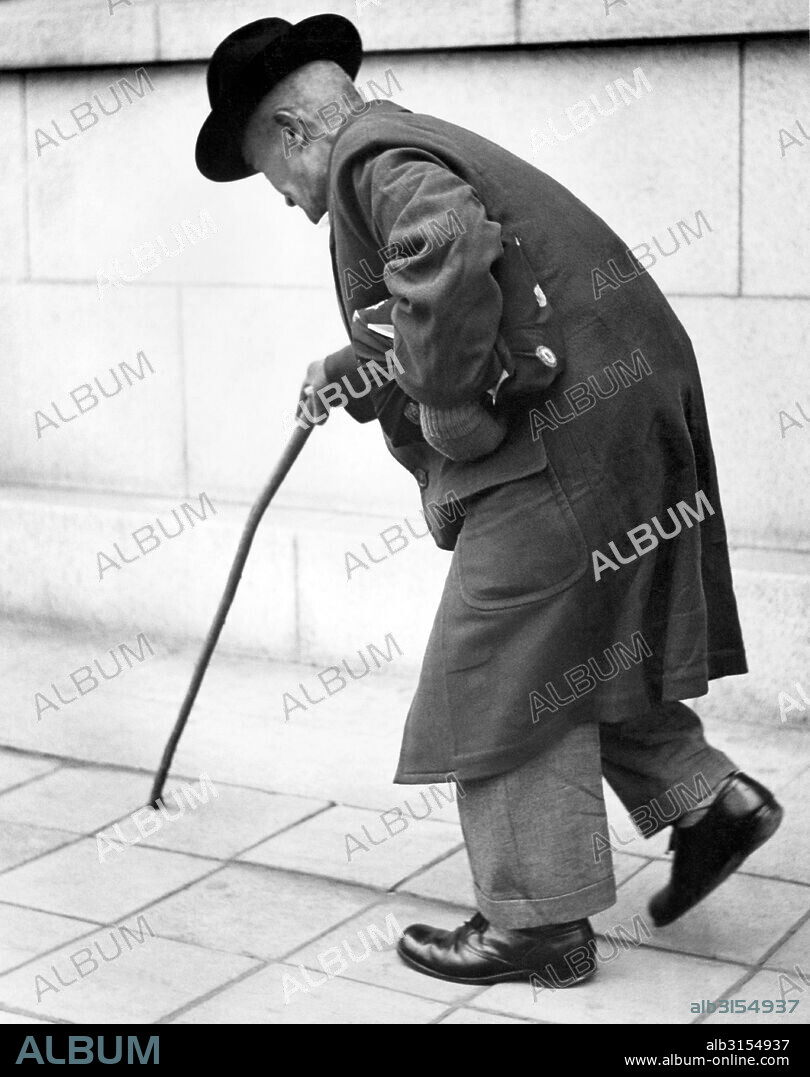San Francisco, California:  c. 1960. A very elderly man walks down a sidewalk using a cane for assistance.