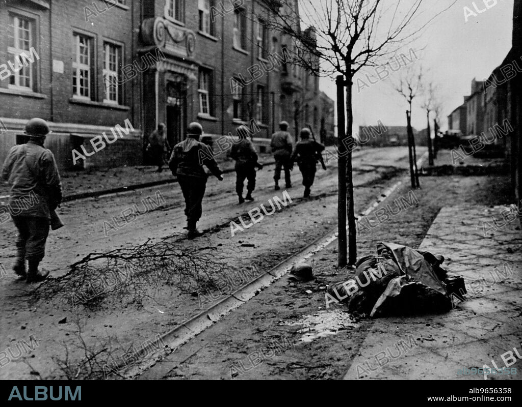 American infantrymen of the 36th, infantry division, 9th. , U.S. Army, file past the body of a dead Nazi. Lying on sidewalk at right, as they move ***** a street in the German Town of Lintfort. March 17, 1945. (U.S Official Photograph).