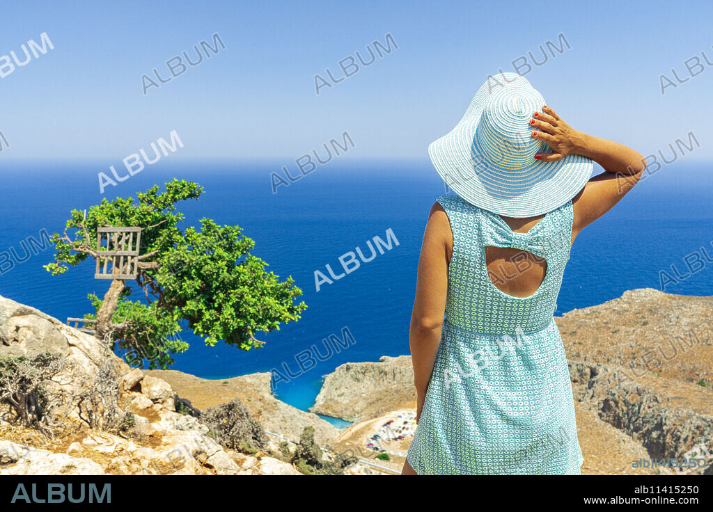Back view of young woman with dress and sun hat watching the sea, Crete, Greek Islands, Greece, Europe.