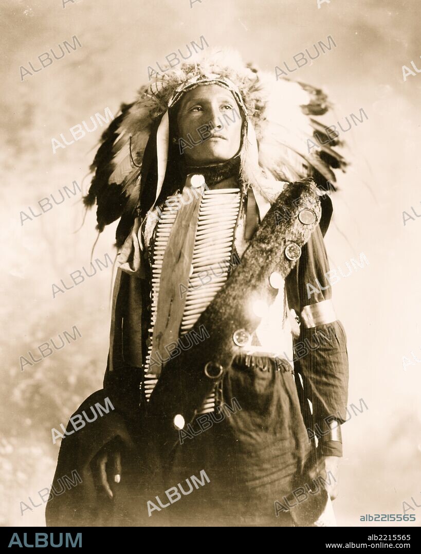 Plenty Holes, a Sioux Indian in feather headdress and bone breast piece, half-length portrait, standing, facing slightly right.