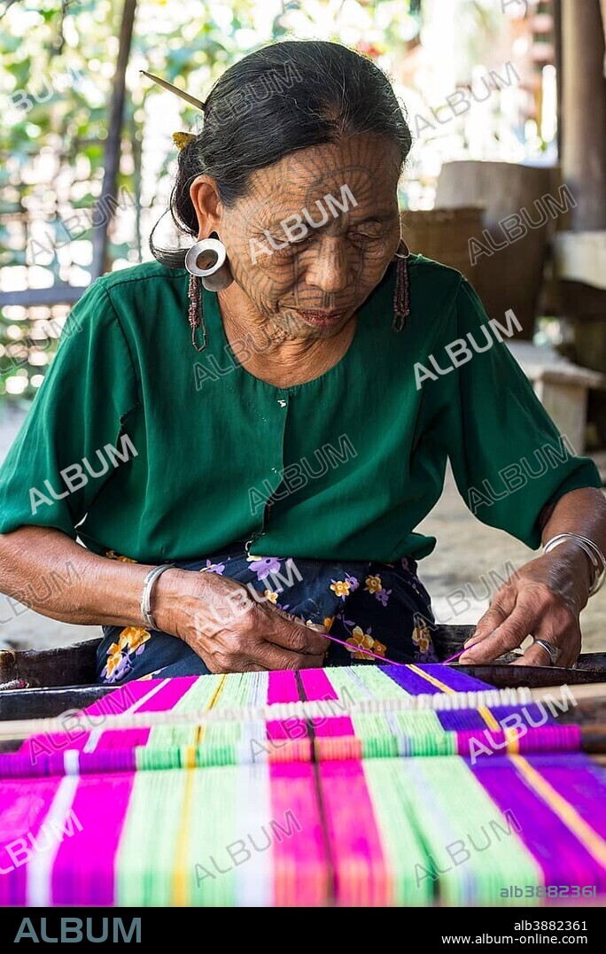 Woman with traditional facial tattoos and ear jewellery from the Chin people, ethnic group, the last of their kind, weaving on a loom, Rakhine State, Myanmar, Asia.