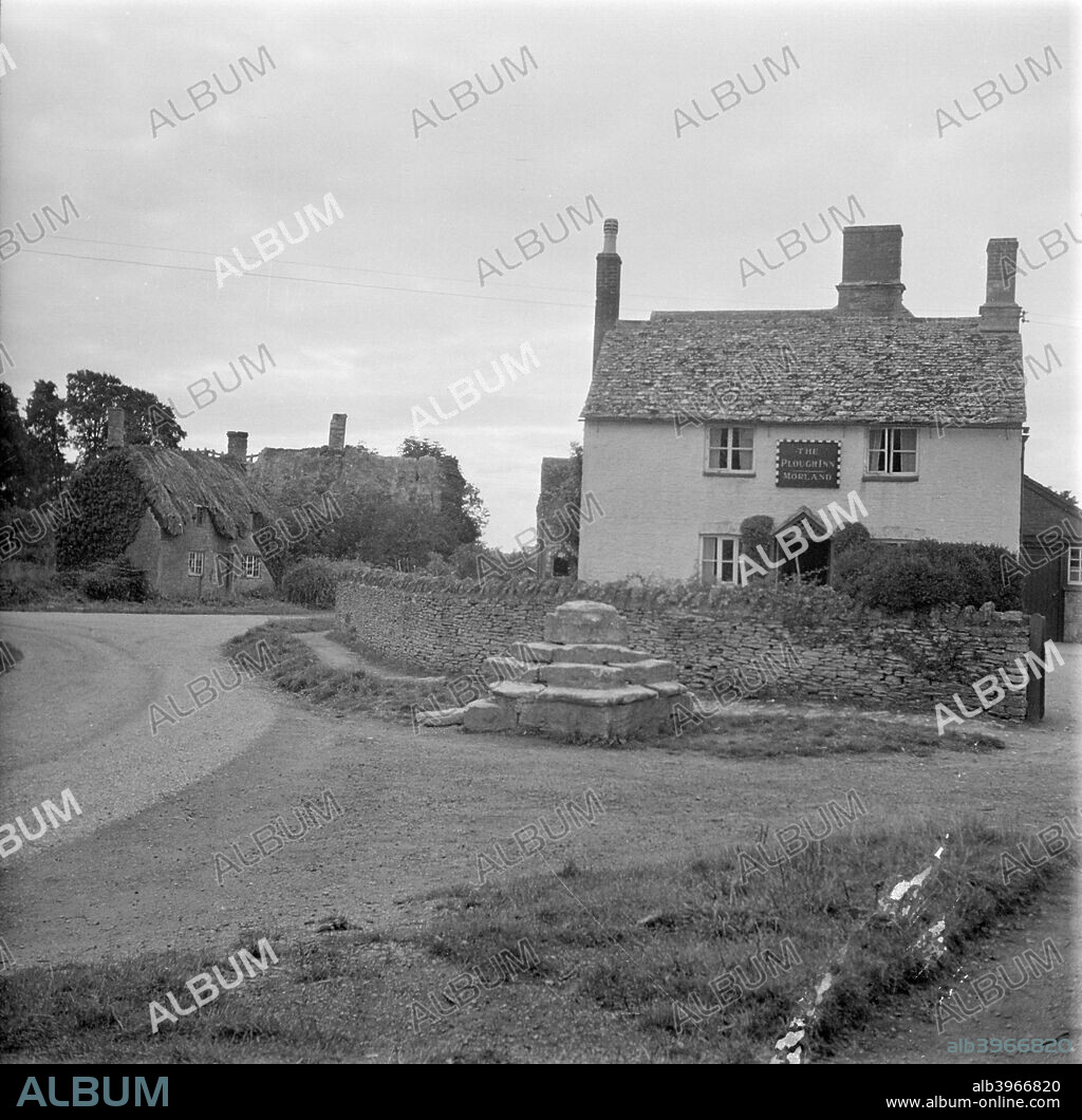 The Plough Inn, Kelmscott, Oxfordshire. The base of a medieval preaching cross stands on the verge in front of the Plough Inn in Kelmscott village. The pub is surrounded by a dry-stone wall. A group of derelict cottages in need of re-thatching can be seen to the left.