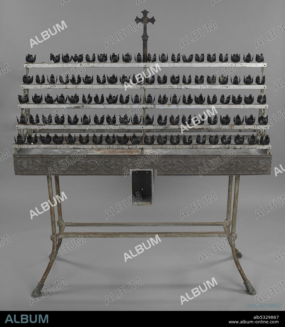 A votive candle stand with ninety-nine candle holders. The holders all feature four stylized leaves. The top of the stand has five rows of with twenty holders each. One of the holders on the top row is missing. The stand is supported by an inverted arch made up of scrollwork with stylized vines on the outside edges and center. The supports are attached to rows of votive holders by screws. The stand is topped with cross. The cross sits on top of a plain column. The cross is capped at the top, bottom, and either arm with trefoils. At the intersection of the arms and panel are two concentric circles with the outside circle appearing behind the cross and the inner circle inside the intersection. Within the inner circle of the intersection is a small Greek cross. The surface of the cross is covered with stylized foliage and leaves.
