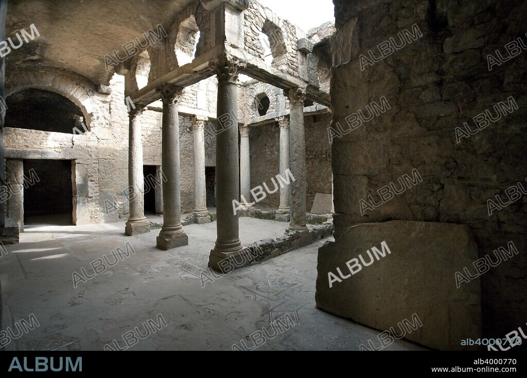 The House of the Hunt, Bulla Regia, Tunisia. The House of the Hunt, which takes its name from a mosaic in the basement, evidently belonged to a wealthy citizen. It was constructed by combining and altering a number of small existing houses. The upper floor is badly ruined, but the basement, lying some 5m (16ft) below ground level, is well preserved. The rooms, laid out round a courtyard lined with Corinthian columns, include bedrooms (identified by the small mosaic-decorated platforms for the beds) and a large dining room (triclinium), also with a pavement mosaic. The bedrooms have an interesting air supply and ventilation system, with pipes built into the walls which communicate with an air shaft surrounding the rooms. The layout on the ground floor is similar, with two colonnaded courtyards, dining and living rooms, bedrooms and a small bath-house. Bulla Regia is an archaeological site in northwestern Tunisia, a former Roman city near modern Jendouba. It is noted for its Hadrianic-era semi-subterranean housing, a protection from the fierce heat and effects of the sun.