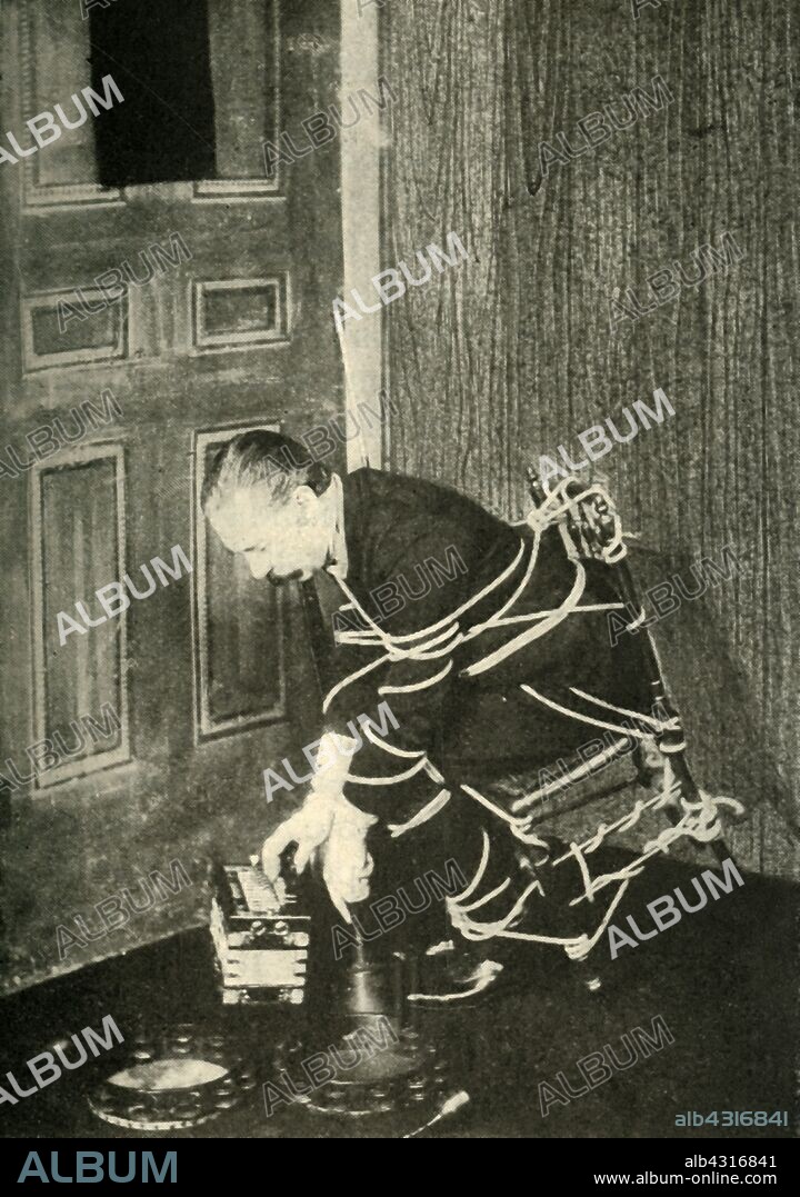 John Nevil Maskelyne performs a 'spirit cabinet illusion', c1910. 'Picking up an accordion and playing it with one hand when tied in the same manner as Mr. Fay'. John Nevil Maskelyne took up a challenge by Sir Hiram Maxim to reproduce a trick originally performed by the Davenport brothers, fraudulent spiritualists from America, and by an imitator of theirs, a Mr Fay. Maskelyne (1839-1917) was a British stage magician, escapologist, paranormal investigator and inventor of the pay toilet. From "Mr. Maskelyne's Reply to Sir Hiram Maxim", published in "The Strand Magazine, an illustrated monthly", Volume XL - July to December 1910. [George Newnes Ltd, London, 1910].