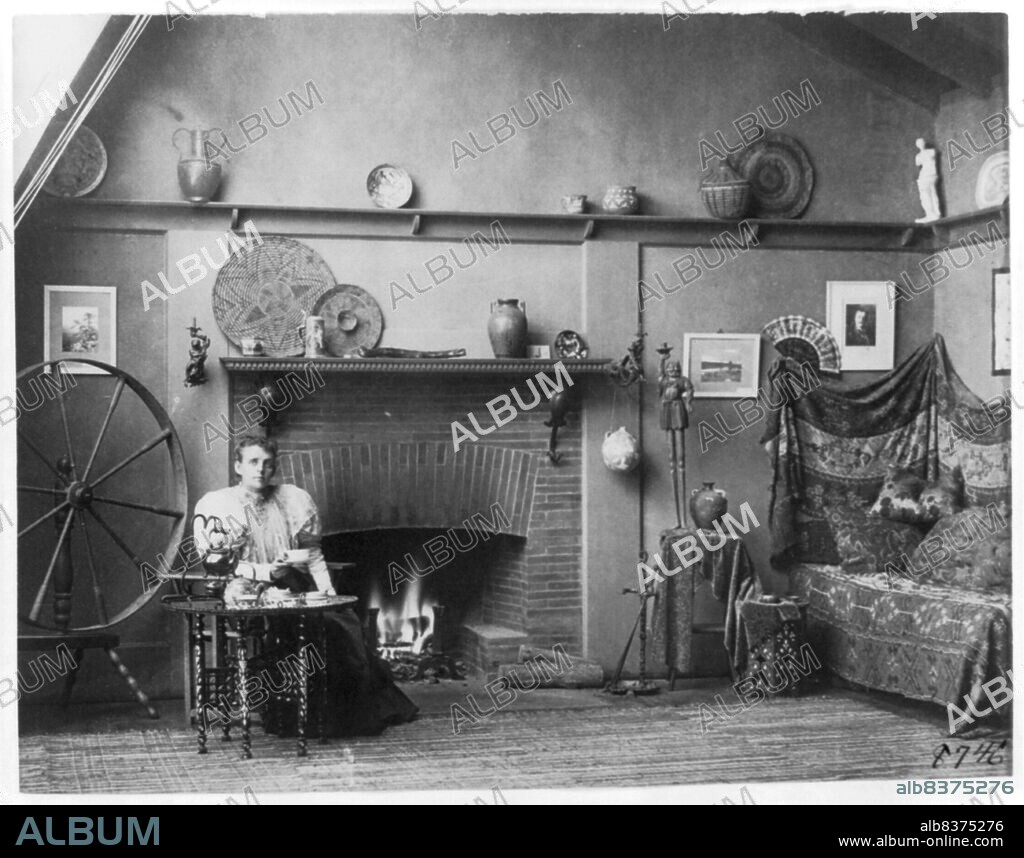 Self-portrait of photographer Frances Benjamin Johnston in her studio at 1332 V Street, N.W., Washington, D.C., c1900. Half length view, seated at small table, facing slightly right; between spinning wheel and fireplace.