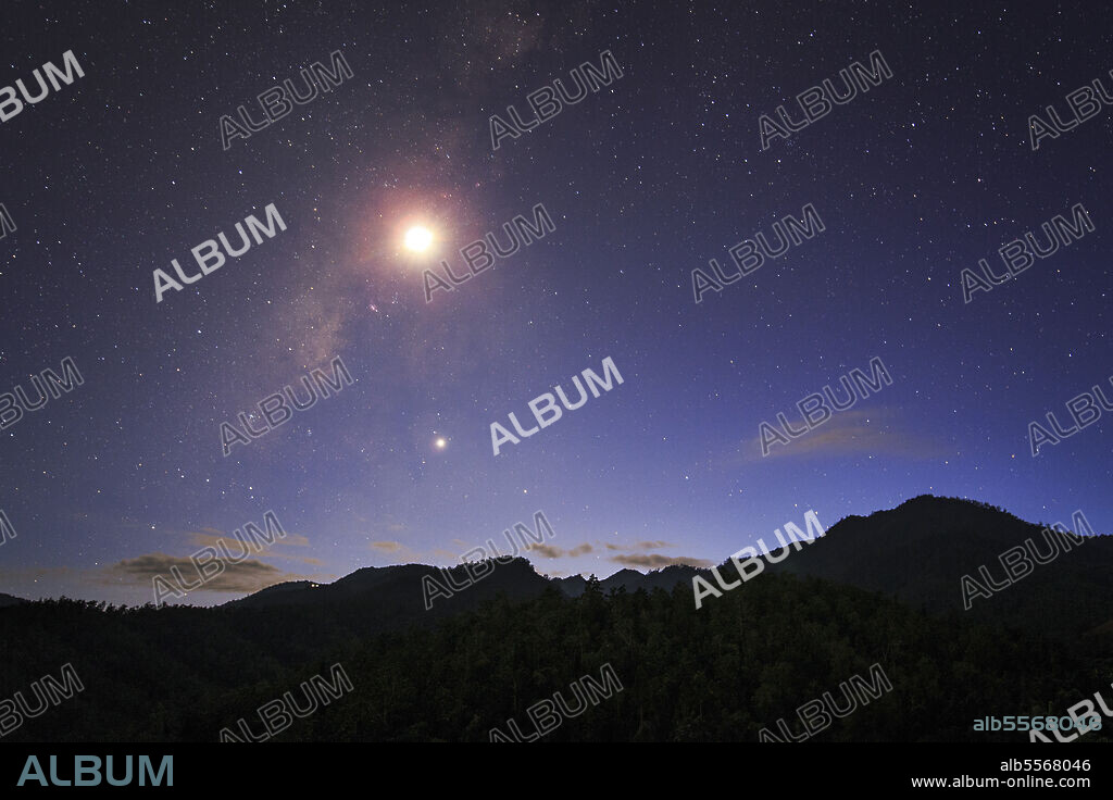 The Milky Way and waxing crescent moon shine above the mountain in Pai, north of Thailand. Venus, the evening star is visible in the evening twilight.