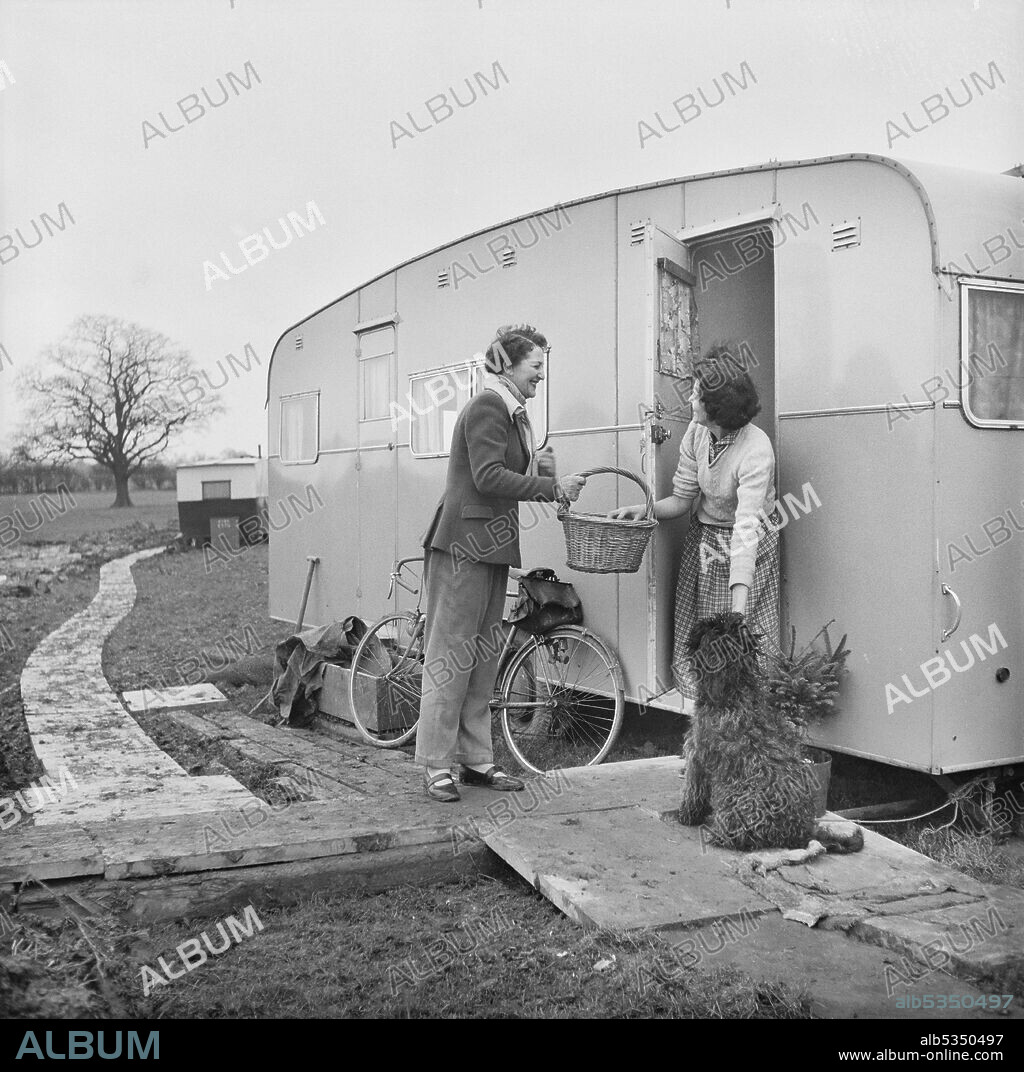 JOHN LAING PLC. The local butcher's wife delivering supplies to Mrs Glover, living in a caravan on a temporary site at Berkeley Nuclear Power Station during its construction. Berkeley Nuclear Power Station was the first commercial nuclear power station in the United Kingdom and one of the first generation of nuclear reactors in Britain. Laing were the civil engineers for the project with work commencing on 7th January 1957. The 57 acre site included offices and temporary housing with facilities for the workers on site. Mrs Glover, shown in the photograph, was the wife of Jim Glover who was a Lima excavator river working on the diversion of the Conigre Pill.