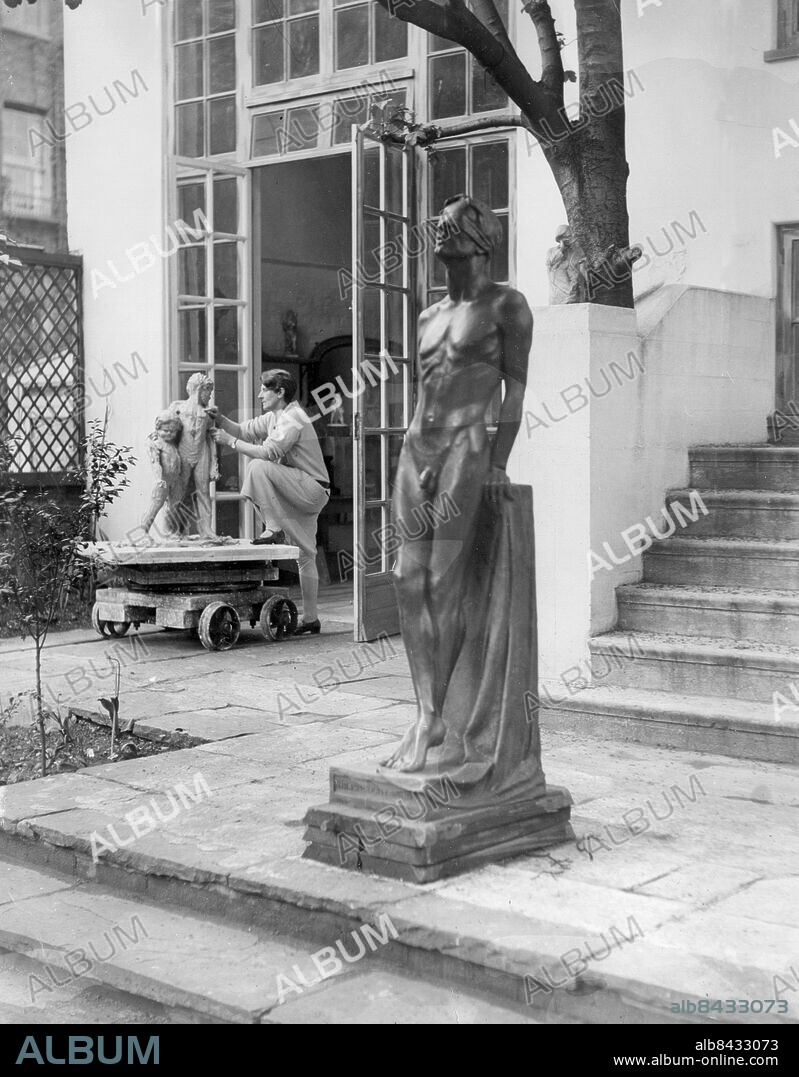 Orig. bildtext... WIDOW OF ANTARCTIC EXPLORER AS A SOCIETY SCULPTRESS. (Exclusive pictures). Lady Hilton Young, who is better known as Lady Scott, the widow of the Antarctic explorer, lives in the quaint cottage, near Kensington Gardens that Sir James Barrie christened The Little White House when he lived in it many years ago. It must be one of London's smallest houses, for it covers barely 30 sq. feet behind Bayswater Rd., but there is one large room where Barrie wrote Peter Pan, and several of his plays. This Lady Young has made her studio.She has fitted a miniature railway between the studio and the back garden so that when the weather is fine she can move her work out into the open on a trolley. In the garden are two of her most charming works, braving the wind and rain. One is the fine statue of a blindfolded youth, which she exhibited in the Academy calling it The Kingdom is Within, and the other is the boy's figure called These Had Most to Give which won a medal at the Paris salon. Photo shows. The statue The Kingdom Is Within, a blindfolded figure of a youth. This is a recent work by Lady Young, who is seen in the background at work in the garden of her home. Anm. K Hilton Young, 1878-1947, baronessan Kennet, född Bruce, brittisk skulptris, änka efter upptäcktsresanden Robert Falcon Scott. Ateljéer Kroppar Ögonbindel CD631 persons: KATHLEEN HILTON YOUNG sites: LONDON;STORBRITANNIEN.