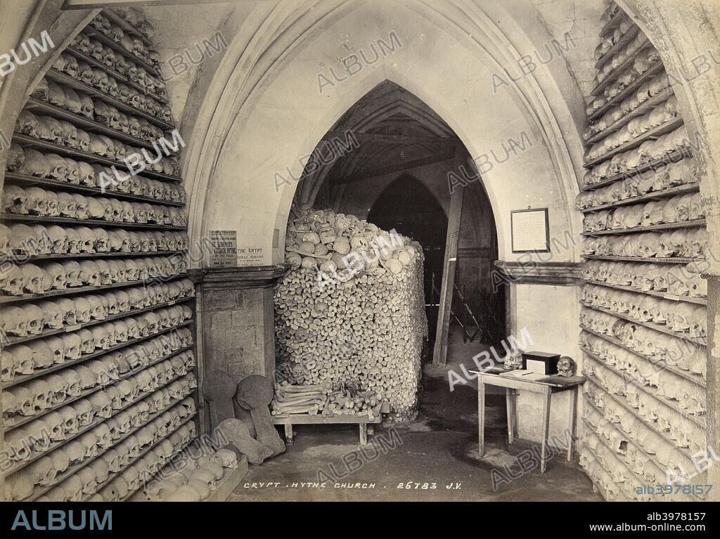 Crypt, St Leonard's Church, Hythe, Kent, c1900. This image shows the ossuary, or bone store, in the vaulted ambulatory beneath the chancel of the church. The space had been used to store skulls and other bones disturbed during building work or when new graves were dug. Some 10,000 bones, including around 2000 skulls, dating from the medieval period were arranged on shelves or stacked in piles. The ossuary had long attracted visitors, and the photograph shows advertisements for guides written by the vicar, and a table with what appears to be a visitors' book and a donations box. Albumen print.