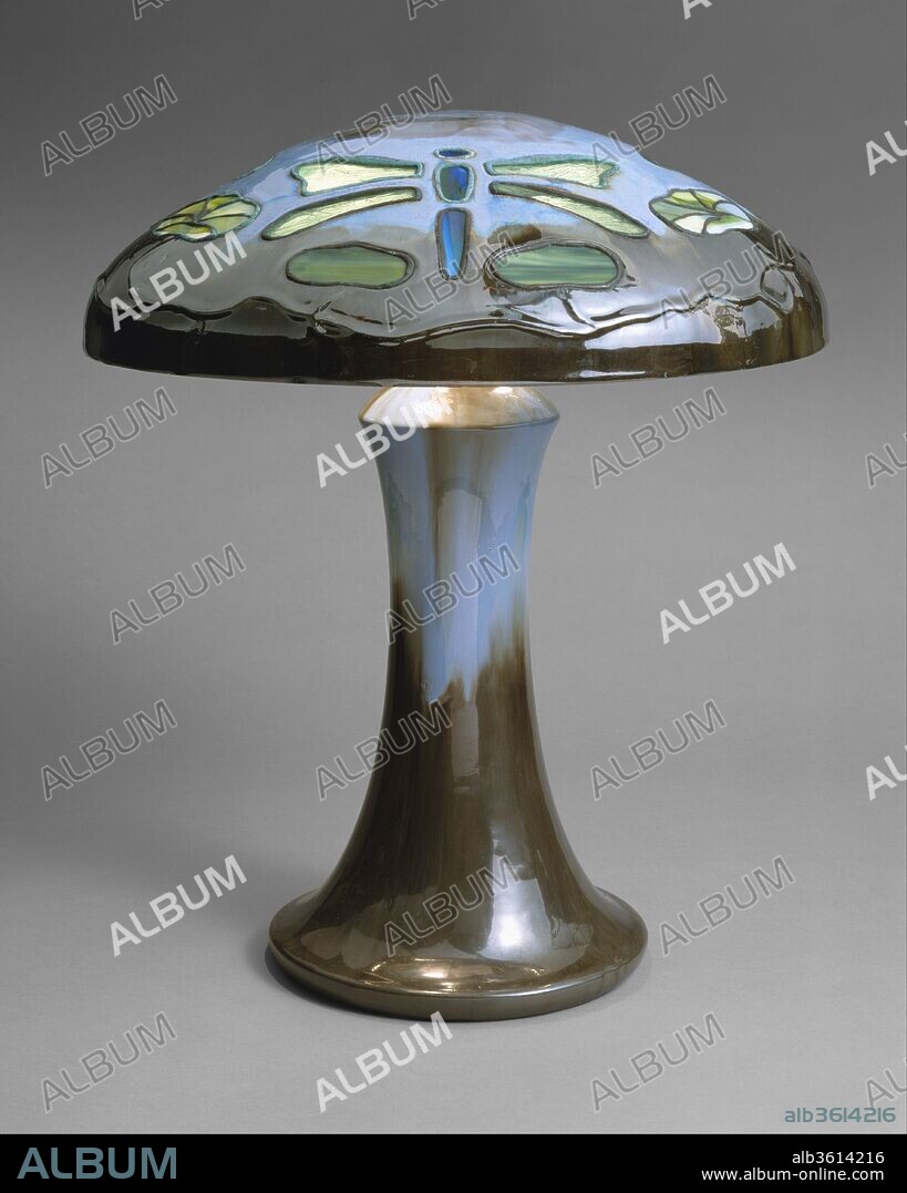 Table Lamp. Artist: Fulper Pottery Company (1899-1935). Culture: American. Dimensions: H. 19 5/8 in. (49.8 cm.); Diam. 17 in. (43.2 cm). Date: 1910-15.
The Fulper Pottery was one of the most prolific, successful, and long-lasting art potteries in America. Fulper had operated in Flemington, New Jersey, producing utilitarian stoneware since the early nineteenth century. It was not until 1909 that the firm developed an artistic line, called "Vasekraft," under the direction of William Hill Fulper II (1872-1928), in whose family the lamp descended. The majority of its production was simple solid oriental shapes with brilliantly colored glazes. Fulper lamps-with glazed pottery shades inset with colored glass-were truly innovative forms. The firm's most spectacular and innovative accomplishments are the table lamps made with glazed pottery bases and shades, which were inset with pieces of colored opalescent glass. This ambitious example, one of only two known of this design, features a shade in the most complex pattern made by the factory. The glass-filled openings delineate dragonflies and water lilies, motifs favored at the same time by noted glass artist Louis Comfort Tiffany. Evoking the natural watery environment of the insects and plants, the lamp is sheathed in a rich Chinese blue flambé glaze.