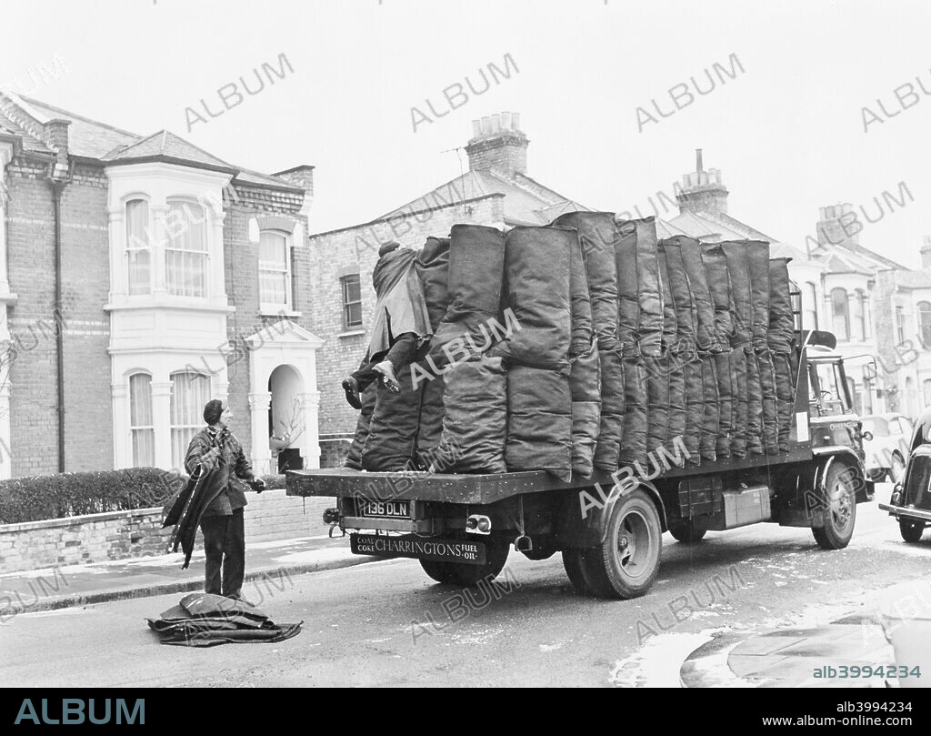Charringtons delivering sacks of coal down a residential street. A coalman watches his colleague reaching over the sacks of coal on the back of the lorry in an attempt to reach something.