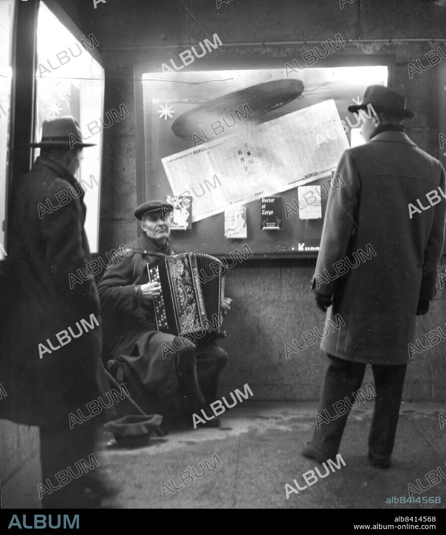 Stockholm 1955-12-31. Medan champagnekorkarna gav fyr på stans femtielva guldkrogar, Olof Widgren läste Nyårsklockan på Skansen och rikets alla domkyrkor ringde in det nya året i radion satt en gammal gatumusikant i trappan mellan Kungsgatan och Regeringsgatan och spelade Stilla natt, heliga natt på sitt illa medfarna dragspel. Foto: Folke Hellberg / DN / TT / Kod: 23. ** SvD OUT **.