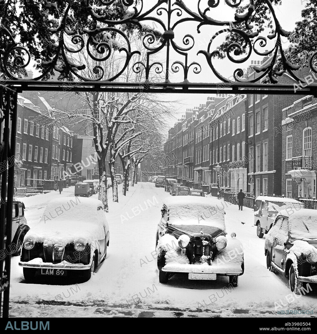 Snow-covered cars, Church Row, Hampstead, London, 1960-1965. A street view in the snow looking east along the middle of Church Row, with a row of cars parked in the foreground and part of the wrought iron overthrow of the church gateway above.