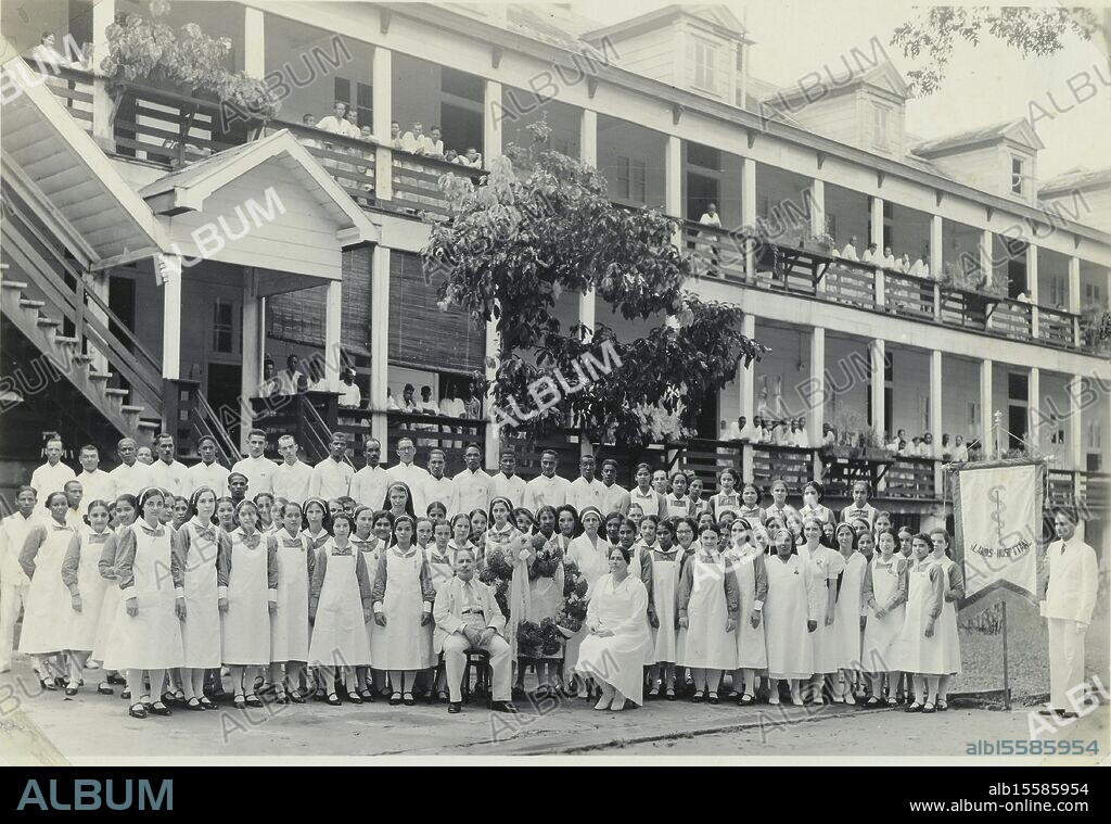 s Lands Hospitaal, Paramaribo, Suriname, Group photo from photo album of hospital staff in front of the building of 's Lands Hospitaal in Paramaribo, Suriname. On the right a man with a banner with the name of the hospital. Part of Theodoor Brouwers' photo album about Suriname around 1930., photograph, anonymous, Paramaribo, 1934 - 1935, paper, gelatin silver print, height, 116 mm × width, 167 mm, height, 148 mm × width, 200 mm.