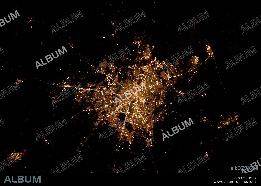 View from space of the city of Houston, Texas at night. The image depicts the roughly 100 kilometer (60 mile) east-west extent of the Houston metropolitan area. Houston proper is at image center, indicated by a "bull's-eye" of elliptical white to orange-lighted beltways and brightly lit white freeways radiating outwards from the central downtown area. Suburban and primarily residential urban land uses are indicated by both reddish-brown and gray-green lighted regions that reflect a higher proportion of tree cover and lower light density. Petroleum refineries along the Houston Ship Channel are identified by densely lit areas of golden yellow light.