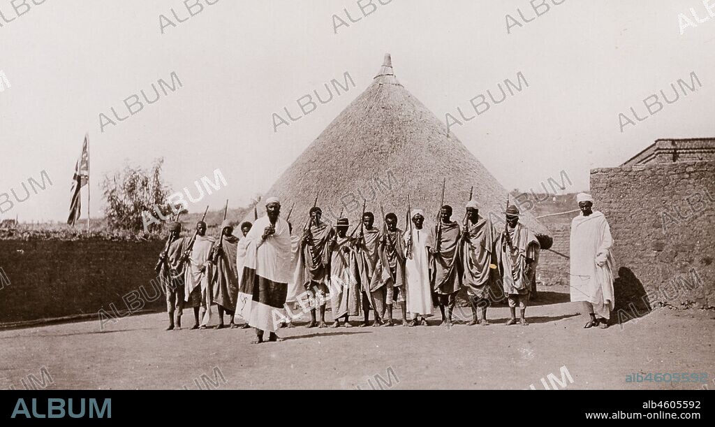 Sudan, Printed caption reads: 'Abyssinians at Gallabat. Published by G N Morhig, The English Pharmacy, Khartoum. Copyright 186'. Armed guard at the Sudan-Ethiopia borderA number of armed Ethiopian men stand in front of a round, thatched hut at Gallabat, guarding the border crossing between Sudan and Ethiopia. A union jack flag is raised on a flagpole to the left of the photograph, [c.1906]. 2003/222/1/2/95.