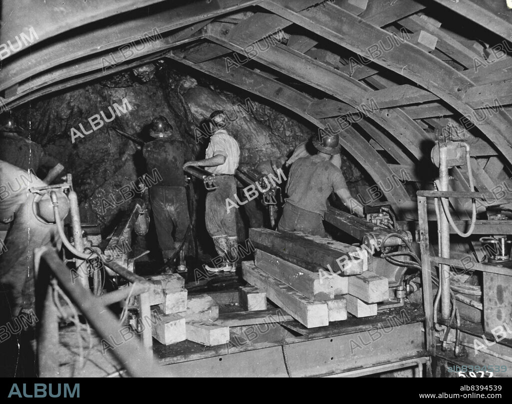 Rock miners working on the top platform of the tunnelling jumbo in the Eucumbene Tumut tunnel. The jumbo has three platforms and a fourth group of men work from the tunnel floor.1949:The Snowy Mountains project was initiated. ***** as one of the biggest civil engineering works in progress ***** in the world. Scheduled for completion by 1975, the Snowy water sources.***** the project cost. April 29, 1955. (Photo by S.M.H.E.A. Official Photograph).