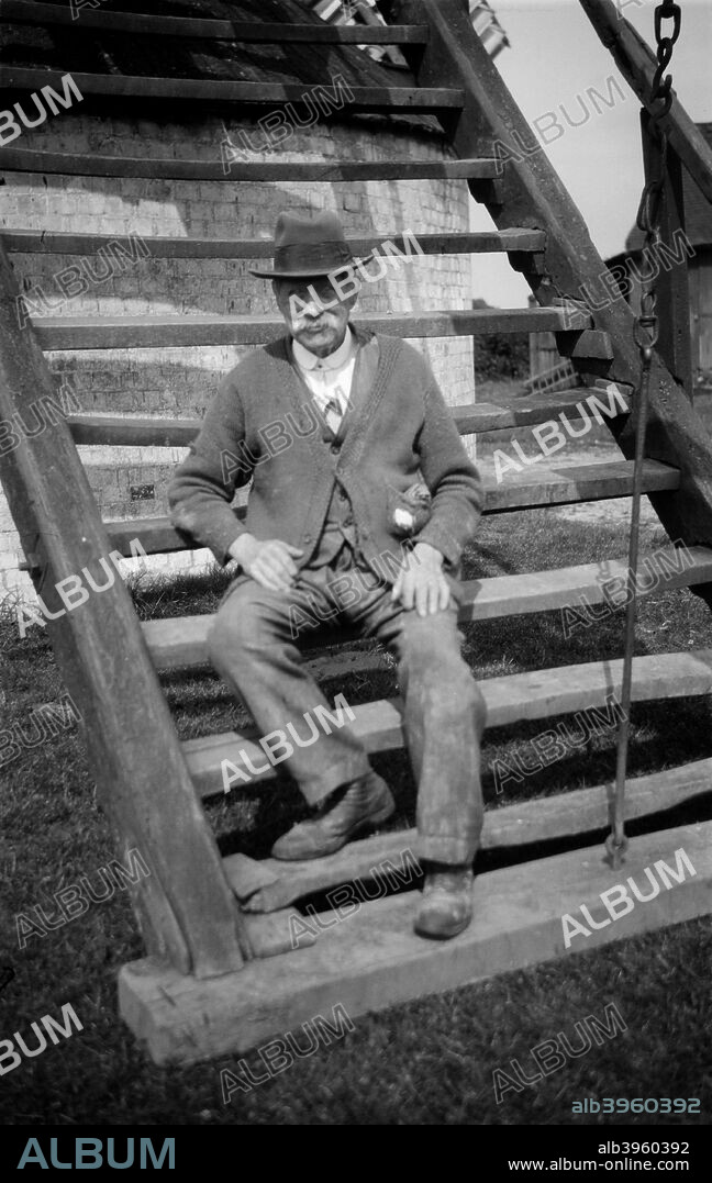 Miller outside his windmill, Outwood, Surrey, 1933. William Jupp, the miller at Outwood, seated on the steps of his post mill. He wears a trilby and a rather worn cardigan.