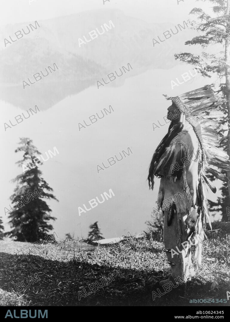 EDWARD SHERIFF CURTIS. Praying to the Spirits at Crater Lake-Klamath, c1923. Klamath Indian chief in feather headdress standing on mountain overlooking Crater Lake, Oregon.