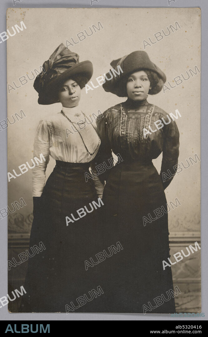 The full length portrait shows the two women standing side by side, wearing large hats with long-sleeved blouses and long skirts. They are looking at the camera with their hands behind their backs.