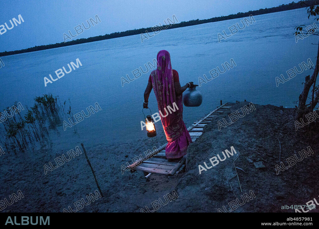 June 22, 2017, West Bengal, India: A woman from Gosaba Island holds a kerosene lamp in one hand and a water jar in the other, as she walks down to the river bank at dusk. Electricity has not reached some of the more remote areas in the Sundarbans. (Credit Image: © Supratim Bhattacharjee/ZUMA Wire).