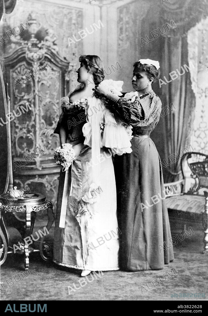 Maid helping woman get dressed in evening gown. Fashion is a popular style or practice, especially in clothing, footwear, accessories, makeup, or furniture. An evening gown or gown is a long flowing women's dress usually worn to a formal affair. Fashion is a distinctive and often habitual trend in the style in which a person dresses. It is the prevailing styles in behavior and the newest creations of textile designers. Prior to the mid-19th century, most clothing was custom-made. It was handmade for individuals, either as home production or on order from dressmakers and tailors. By the beginning of the 20th century, with the rise of new technologies such as the sewing machine, the development of the factory system of production, and the emergence of department stores, clothing had increasingly come to be mass-produced in standard sizes and sold at fixed prices.