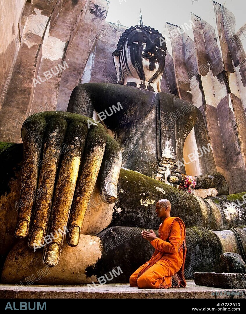 Praying monk neeling in front of the hand of the Buddha statue Phra Achana which is decorated with gold leaf, Wat Si Chum, Sukhothai Historical Park, Sukhothai, Sukhothai Province, Northern Thailand, Thailand, Asia.