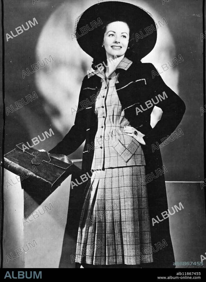 Margaret Lockwood, glittering young star of the J. Arthur Rank organisation, takes on a military mien in this guardsman-inspired coat in navy wool. The short-sleeved suit, in grey,navy and red Tweed, has a flared skirt and the jacket is gathered at the waist, accentuated the hipline. Joy Ricardo has created the stunning ensemble for the First Lady of British films. 24 December 1947.