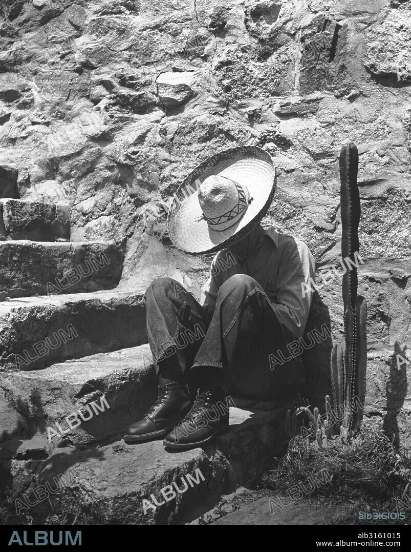 c. 1950 A man wearing a sombrero takes a siesta on steps next to a cactus.