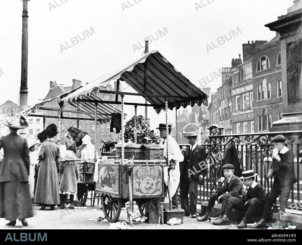 Ice cream sellers, Market Place, Nottingham, Nottinghamshire, c1910. The stall in the foreground belongs to Solari's, who by the sound of the name were probably Italian, as most ice cream manufacturers-sellers were. The people leaning up against the railings of Queen Victoria's statue are eating ice cream from a 'poke' (which supplied the common name for ice cream - Hokey-Pokey, and led to the rhyme which grandmothers were fond of reciting: Hokey-Pokey penny a lump, the more you eat the more you trump!).The picture also shows shops on Beastmarket Hill in the background.