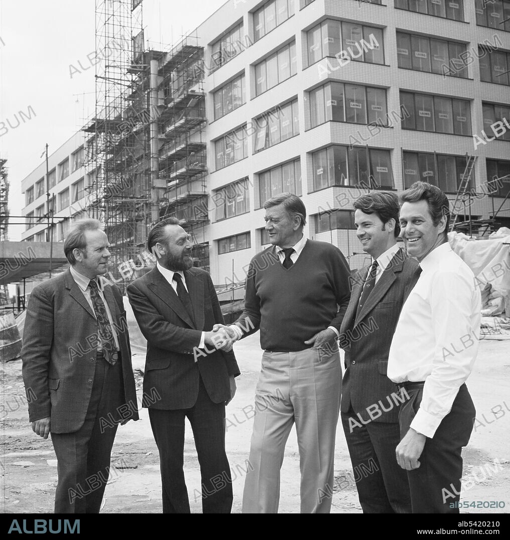 JOHN LAING PLC. The actor John Wayne meeting staff working at the St Thomas' Hospital construction site. This photograph was taken during the filming of Brannigan, directed by Richard Attenborough. Shots were filmed at St Thomas' Hospital and John Wayne along with his co-star, Judy Geeson, and the director were getting to know the site. The photograph was also published in September 1974 in the Laing monthly newsletter 'Team Spirit'. It shows various members of staff who worked on the Laing building contract at the hospital and John Wayne is seen shaking the hand of the projects manager, D Upton.