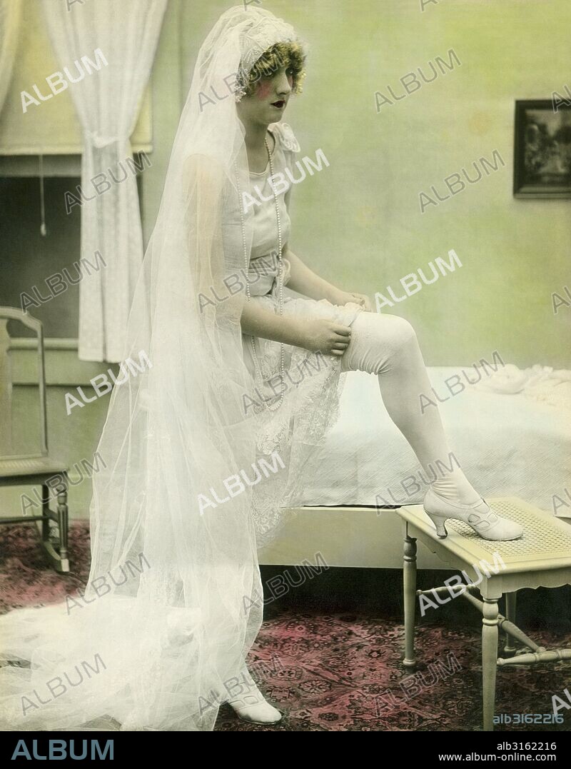 United States: c. 1925. A hand tinted photograph of a nervous looking bride taking off her stockings and titled, "The Bride Retires".
