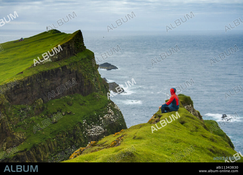 Man sitting on cliffs looks towards the lighthouse, Mykines island, Faroe Islands, Denmark, Europe.