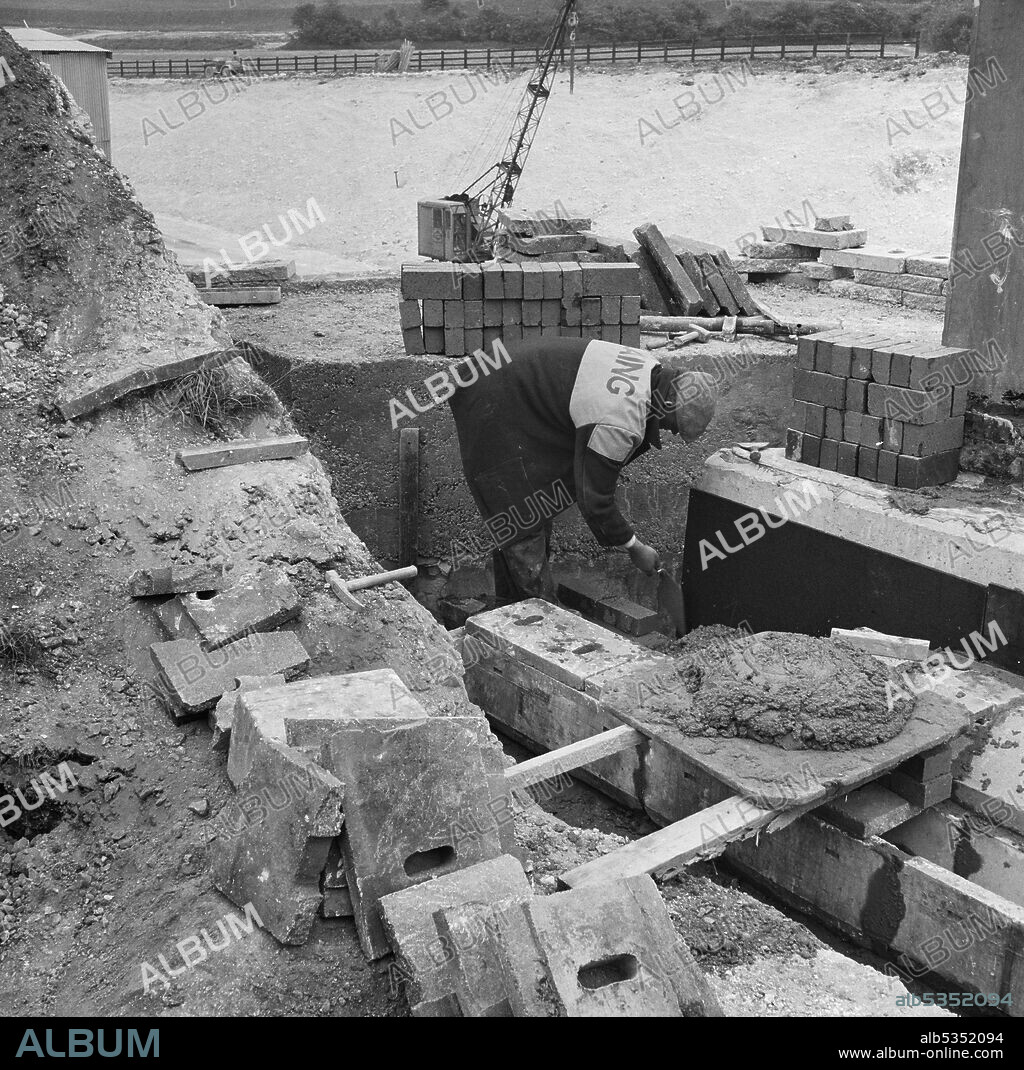 JOHN LAING PLC. A Laing labourer laying bricks near a drainage channel during the construction of Structure 9, a bridge carrying the Luton-Dunstable railway line over the London to Yorkshire Motorway (the M1). The caption beneath the corresponding album print states that this is 'blue brickwork'.
