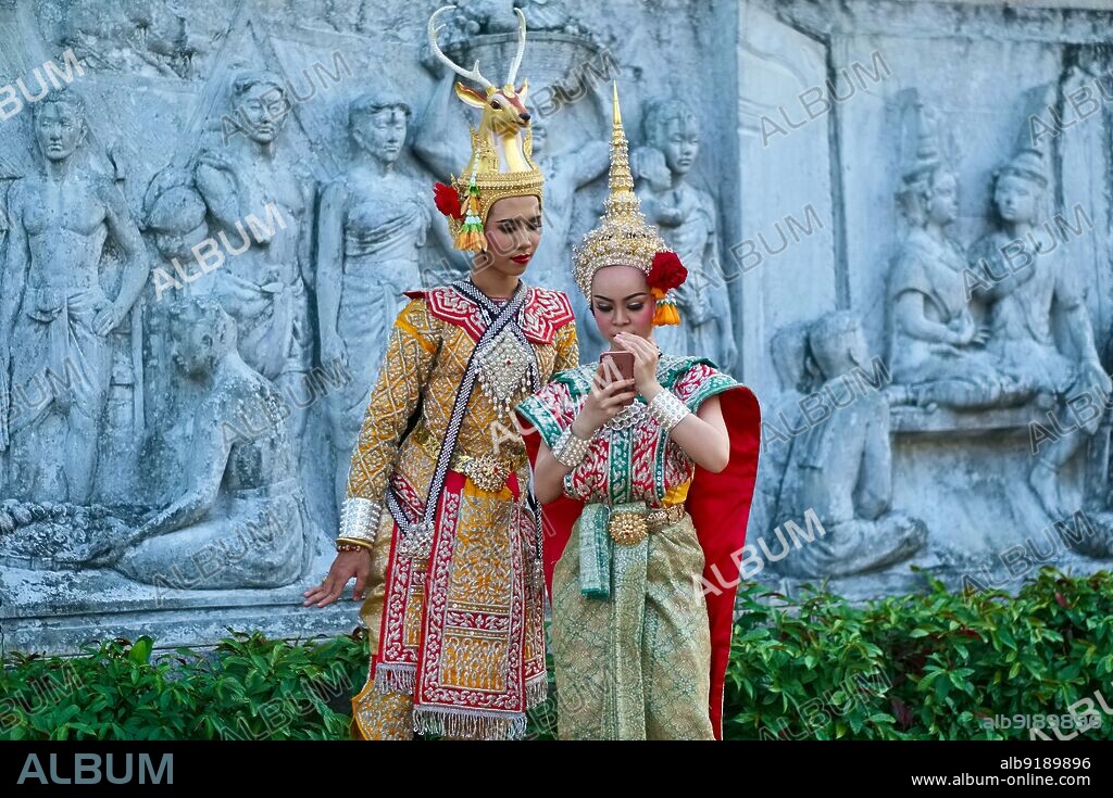 Thailand: Members of a <i>lakhon</i> (traditional Thai drama) troupe during a performance break check their social media accounts; the background shows a frieze depicting historical scenes. Santichaiprakarn Park, Banglamphoo, Bangkok.. <i>Lakhon nai</i> is a performing art originating in the royal court of Thailand (formerly Siam). It features a slow choreography accompanied by a <i>piphat</i> ensemble (traditional musical group). A variation of this genre with male performers is called <i>lakhon nai phu chai</i>.