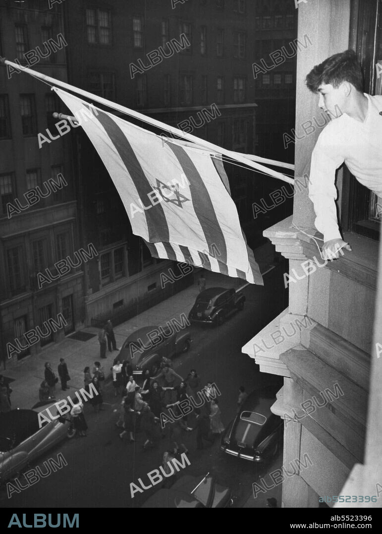 Dance in Street as Palestine Flag Flies - Haim Shertok, 14-year-old son of Moshe Shertok, Foreign Minister of the Newly-Proclaimed Jewish State, Looks out of Window of Jewish Agency at 16 east 66th street, New York City, May 14, as young Jews Dance in the street to celebrate the raising of the Zionist Flag next to the American Flag in Window of Agency. Young Shertok assisted in Ceremony at which flag was flown. The Young Jews in street are dancing the Palestinian Folk Dance, "The Hora." May 14, 1948. (Photo by Associated Press Photo).