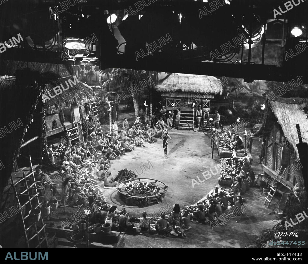 ***** Native in Hollywood -- The still photographer perches ***** on a sound stage catwalk to snap this picture of the filming of a jungle feast scene in Universal - International's Technicolor adventure film, "East of Sumatra". The girl performing the Sumatran dance in Suzan Ball. Films General. February 22, 1954.